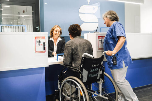 Das Bild zeigt eine Mitarbeiterin, welche eine im Rollstuhl sitzende Patientin zu einem Termin begleitet. 