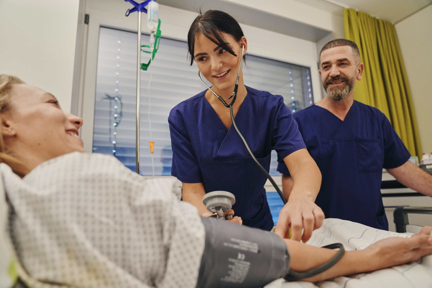 Das Bild zeigt zwei Pflegefachkräfte bei der Untersuchung einer im Bett liegenden Patientin.