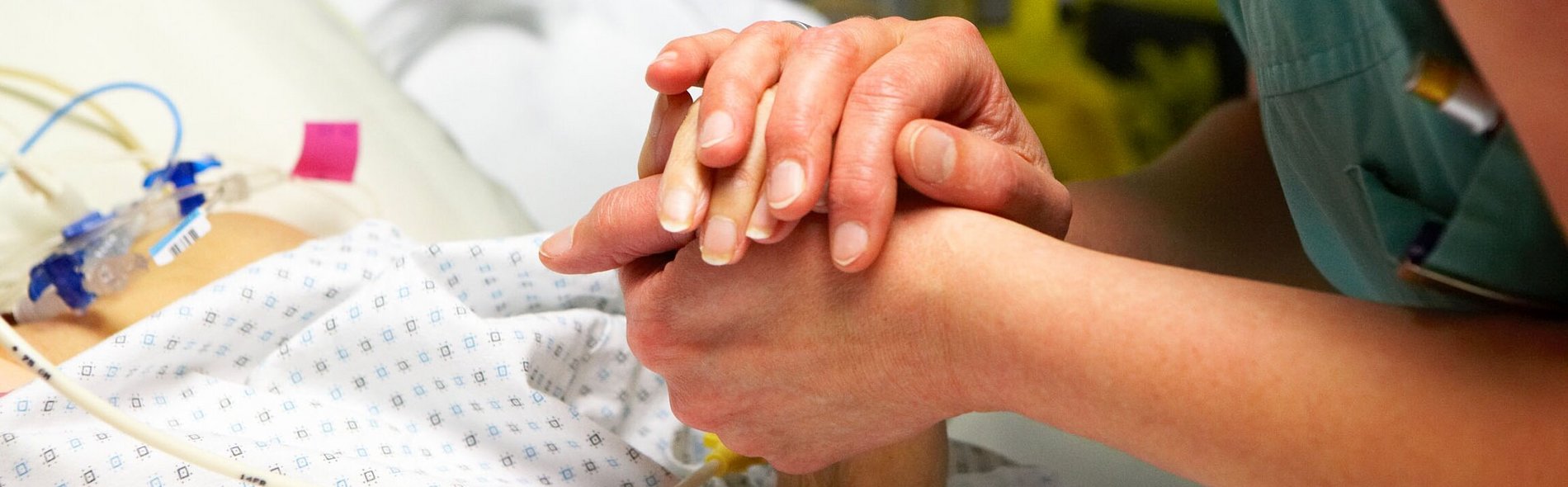 Eine Pflegekraft hält in einem Krankenhaus voller medizinischer Geräte liebevoll die Hand einer Patientin und vermittelt ihr damit Fürsorge und Unterstützung.