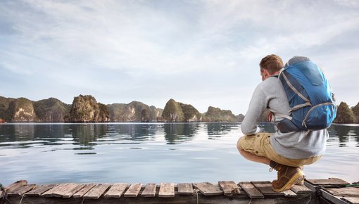 Junger Mann mit einem Rucksack hockt an einem Holzsteg vor Wasser und fernen Bergen.