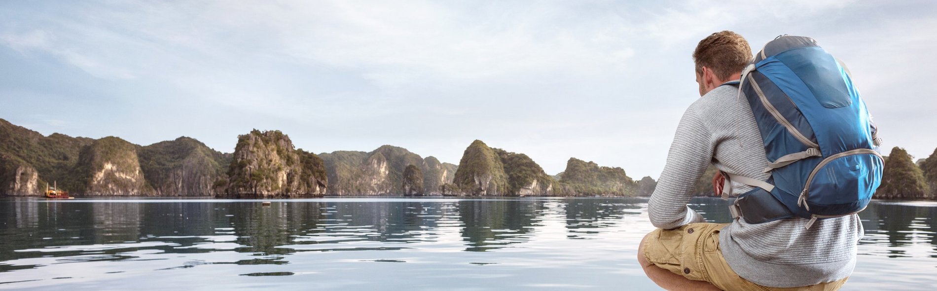Junger Mann mit einem Rucksack hockt an einem Holzsteg vor Wasser und fernen Bergen.