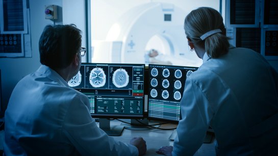 Two neurology physicians examine brain scans on the screen. Patient in MRI can be seen in the background.