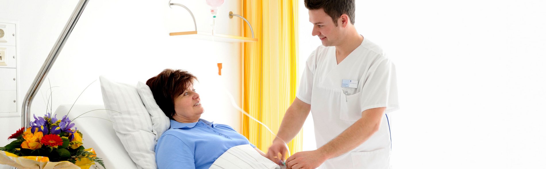 Ein Krankenpfleger in einem weißen Kasack kümmert sich um einen Patientin in einem Krankenhausbett und richtet ihre Decke. Der Raum ist hell, mit gelben Vorhängen und einer Vase mit bunten Blumen auf einem Tisch, was eine fürsorgliche und ruhige Atmosphäre vermittelt.