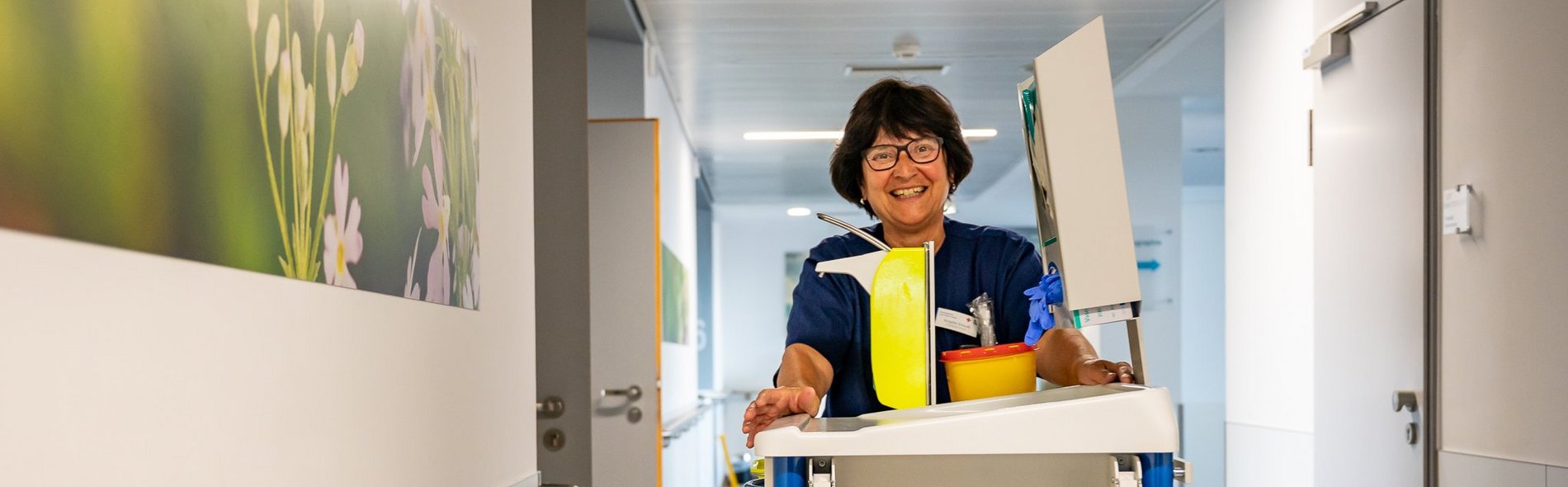 Lächelnde Pflegefachkraft mit Pflegewagen auf dem Weg zum Patienten im modernen Flur der Wahlleistungsstation