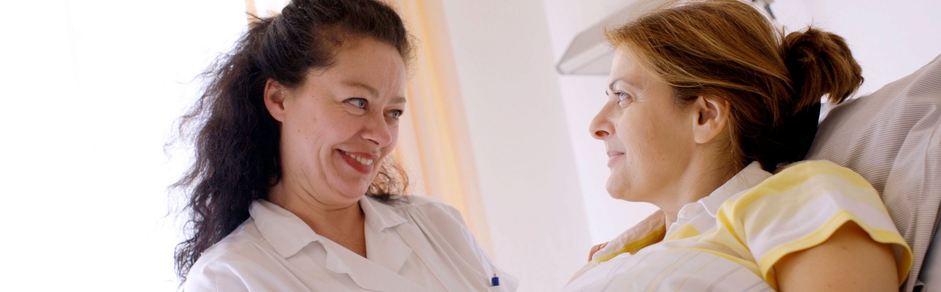 Eine Krankenschwester in weißer Uniform lächelt warmherzig und hält die Hand einer Patientin in einem Krankenhausbett. Die Szene vermittelt Fürsorge und Mitgefühl in einem beruhigenden, sanft beleuchteten Raum.