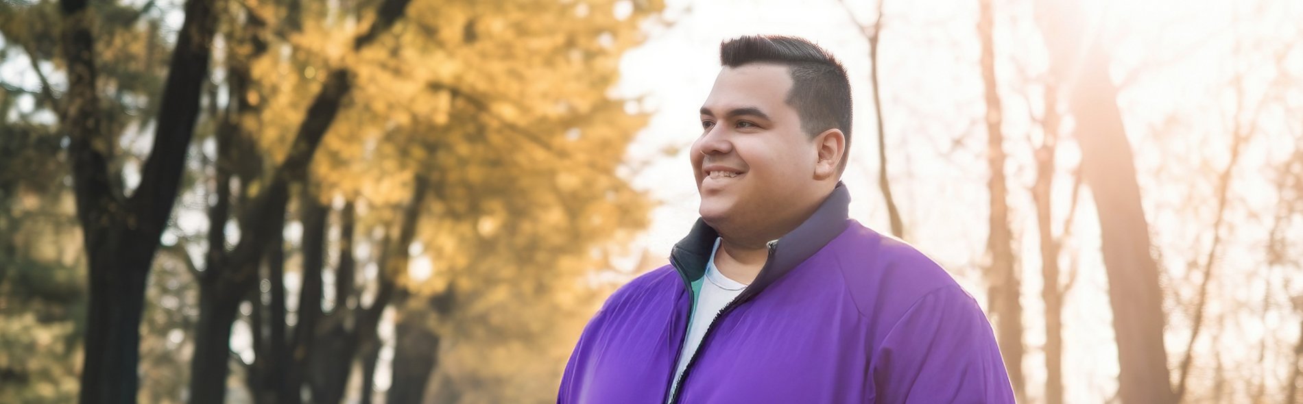 Mann mit lila Jacke steht mit den Händen in den Taschen auf einem von Bäumen gesäumten Weg im Gegenlicht