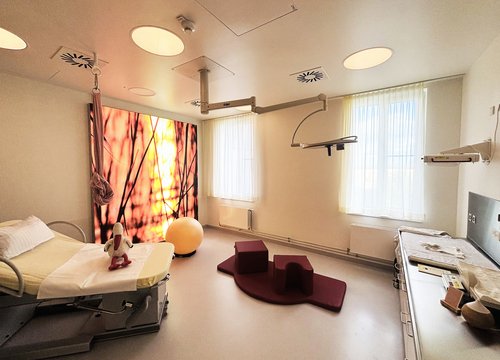 Kreißsaal 1 im Geburtszentrum Biberach, in rot gehalten, mit Entbindungsbett, Be-up Modulen sowie einem großen beleuchteten Panoramabild in rot und orange