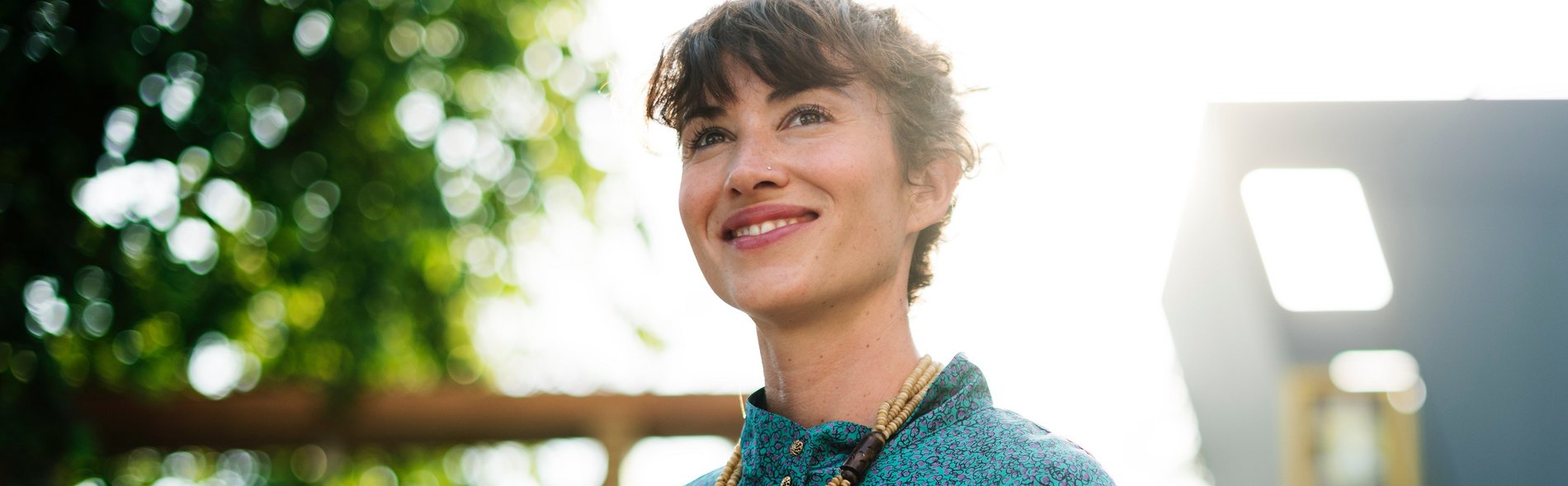 Eine Frau in einem blaugrünen Shirt lächelt, während sie im Freien ein Smartphone in der Hand hält. Sonnenlicht fällt durch die Bäume und schafft eine warme und optimistische Atmosphäre.