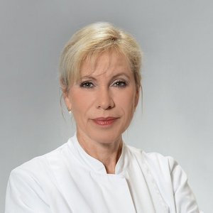 Portrait einer blonden Frau in weißem Kittel mit verschränkten Armen und fokussiertem Blick