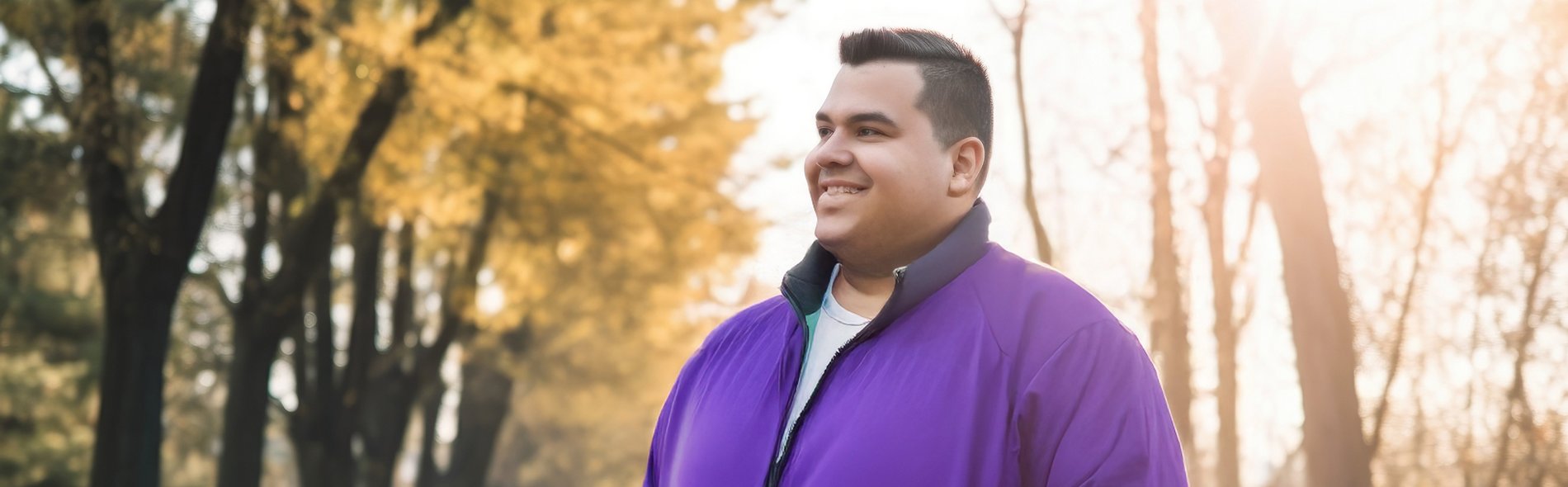 Mann mit lila Jacke steht mit den Händen in den Taschen auf einem von Bäumen gesäumten Weg im Gegenlicht