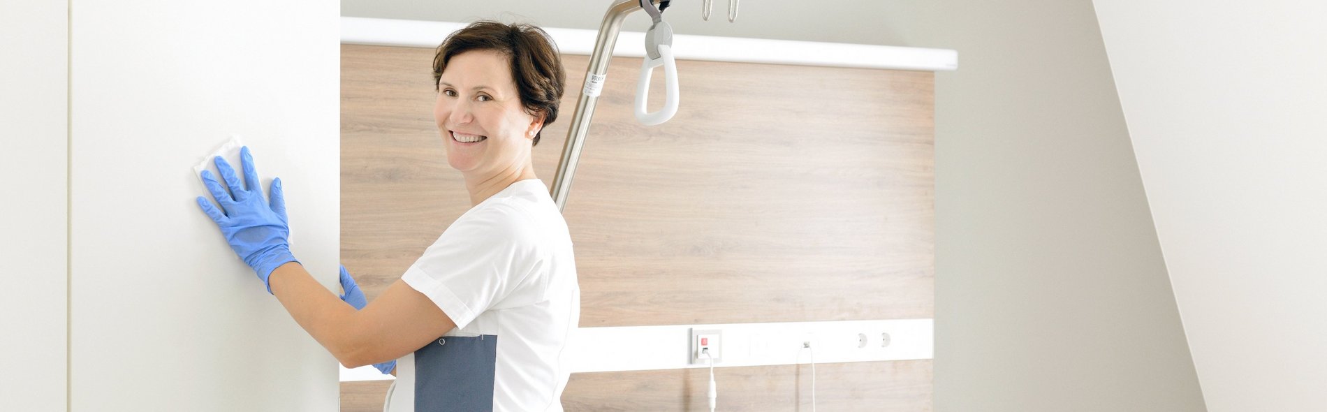 Eine lächelnde Person mit blauen Handschuhen reinigt ein modernes Krankenhauszimmer. Ein ordentlich gemachtes Bett, ein Nachttisch mit Getränken und eine helle Beleuchtung vermitteln eine saubere, einladende Atmosphäre.