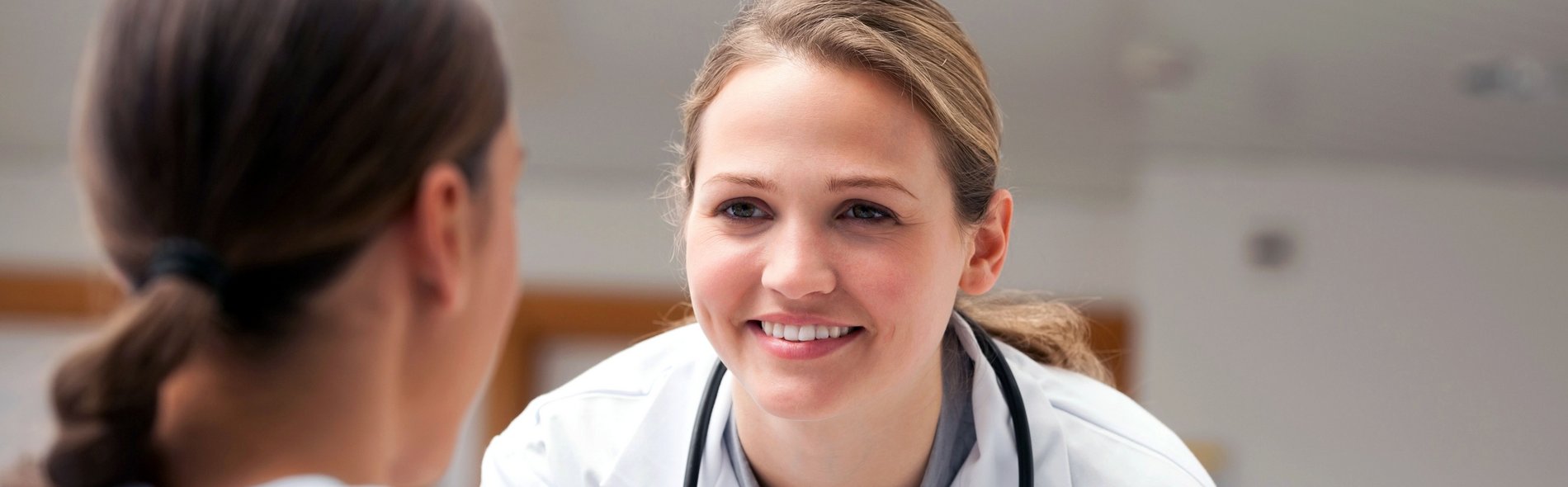 Eine lächelnde Ärztin in einem weißen Kittel mit einem Stethoskop, die sich herzlich mit einer sitzenden Patientin unterhält und dabei Fürsorge und Professionalität vermittelt.