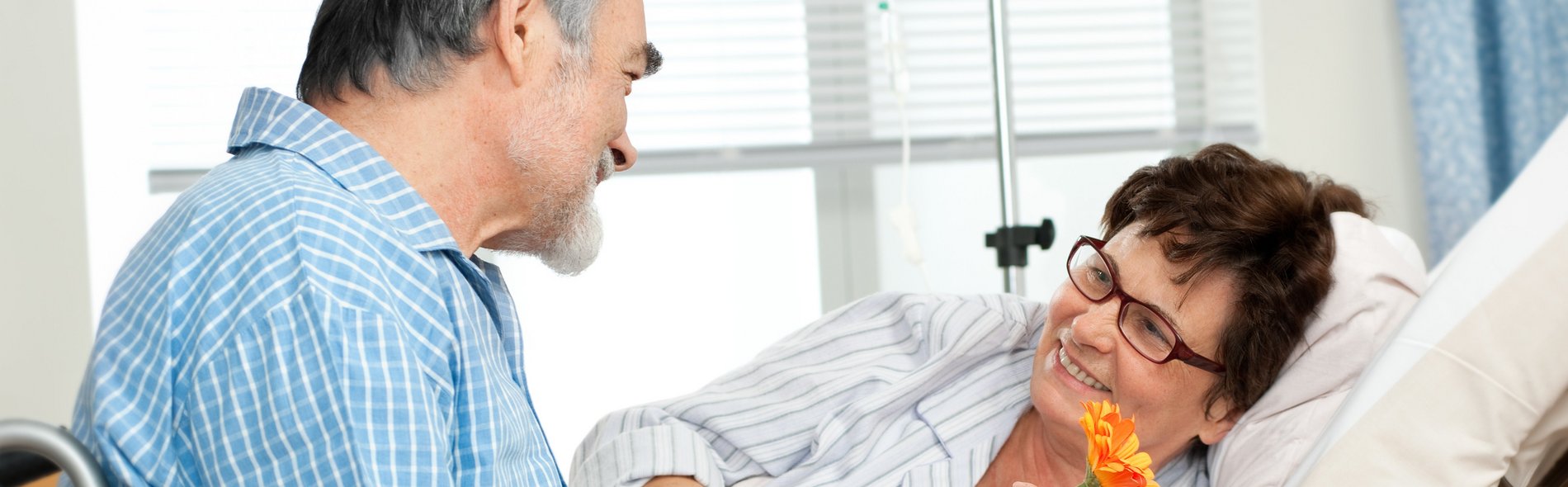 Eine Patientin freut sich über den Besuch ihres Angehörigen. Ein älterer Mann in einem blauen Hemd lächelt eine Frau mit Brille im Krankenbett, die einen gestreiften Pyjama trägt und eine leuchtend orangefarbene Blume in der Hand hält, warmherzig an und vermittelt dabei Wärme und Fürsorge.