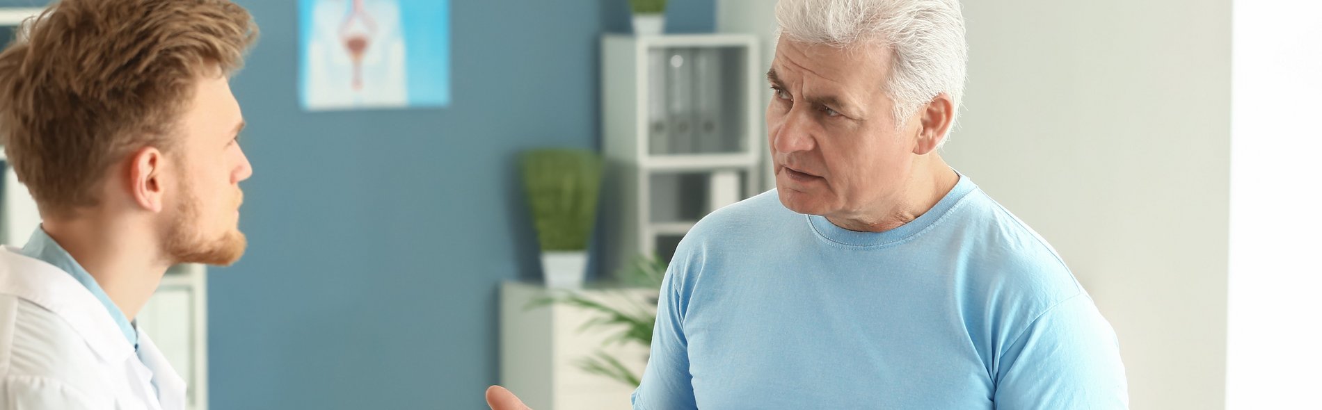 Ein älterer Mann in einem blauen Hemd konsultiert einen Arzt in einem weißen Kittel, der in einem hellen Untersuchungszimmer sitzt.