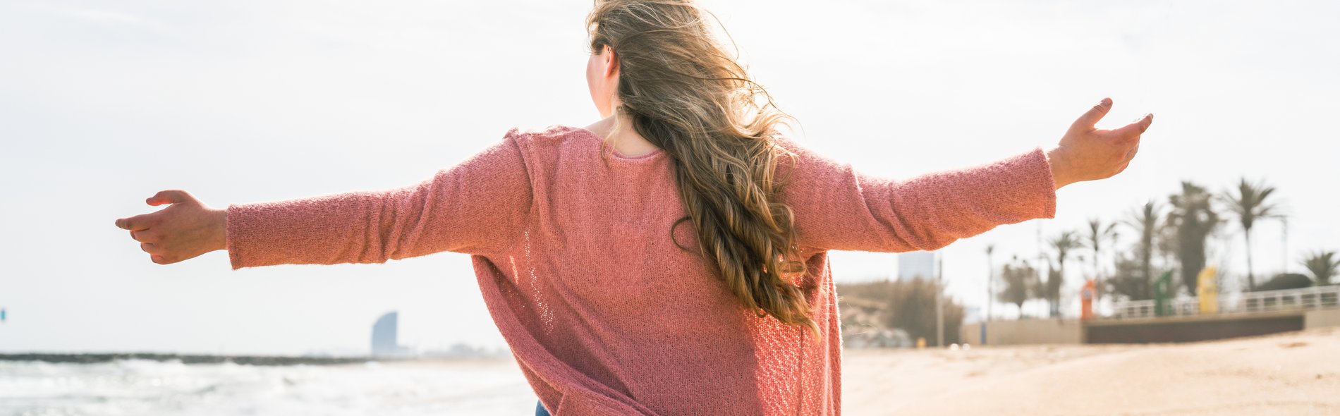 Frau mit langem Haar und offenem rosa Pullover steht mit ausgebreiteten Armen am Strand und blickt aufs Meer