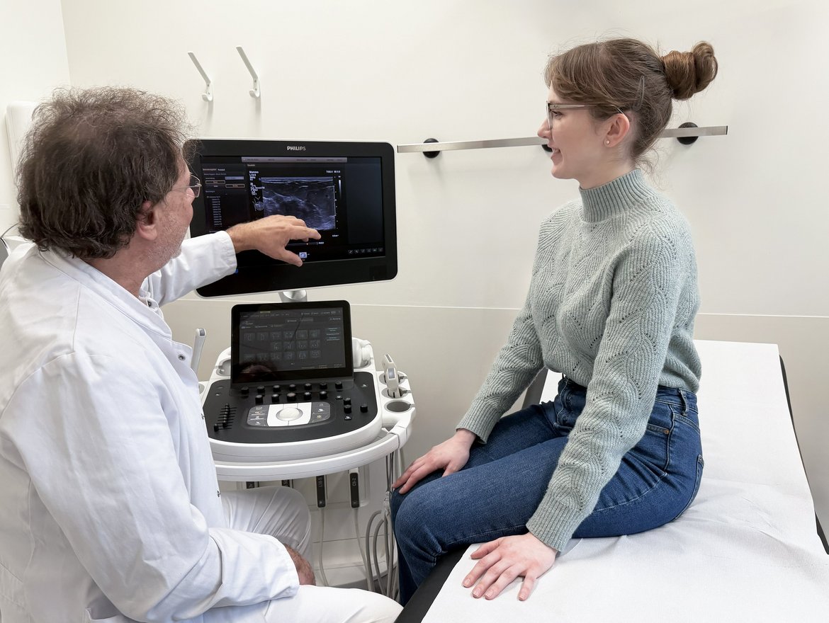 Ultraschall Brust Dr. Steffen Fritz zeigt auf den Monitor des Ultraschallgeräts und erklärt die Aufnahme seiner Patientin, die auf der Liege neben ihm sitzt