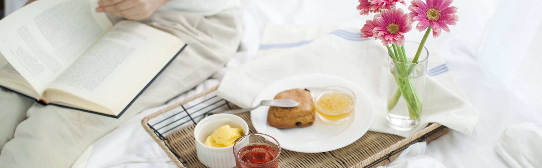 Eine Person in Weiß sitzt auf einem Bett und liest ein Buch. Ein Frühstückstablett mit rotem Tee, einem Scone, Butter und rosa Blumen sorgt für eine ruhige, gemütliche Atmosphäre.