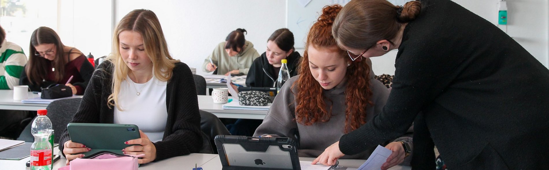 Blick in einen Klassenraum, Auszubildende arbeiten mit Tablet und Schreibblock, Lehrerin gibt Hilfestellung.