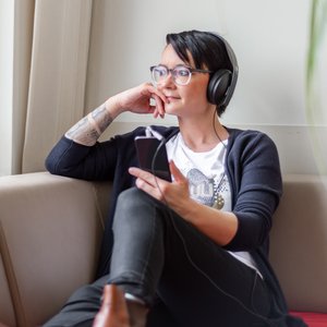 Junge Frau mit Kopfhörern hört auf dem Sofa sitzend Musik oder einen Podcast und sieht dabei aus dem Fenster