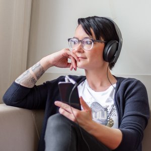 Junge Frau mit Kopfhörern hört auf dem Sofa sitzend Musik oder einen Podcast und sieht dabei aus dem Fenster