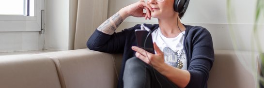 Junge Frau mit Kopfhörern hört auf dem Sofa sitzend Musik oder einen Podcast und sieht dabei aus dem Fenster