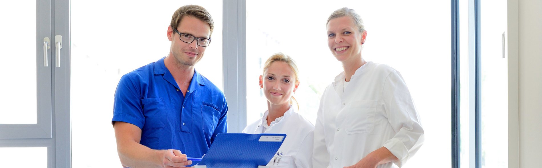 Drei medizinische Fachkräfte in weißen Kitteln stehen in einem hellen Raum. Eine von ihnen hält eine blaue Mappe in der Hand und lächelt. Hinter ihnen sind große Fenster zu sehen.
