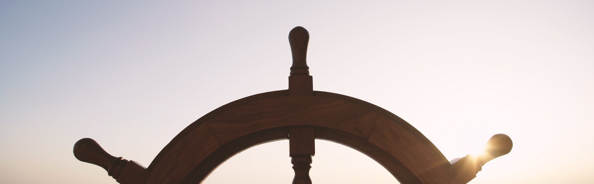Steuerrad eines alten Segelschiffs im Vordergrund während im Hintergrund die Sonne untergeht