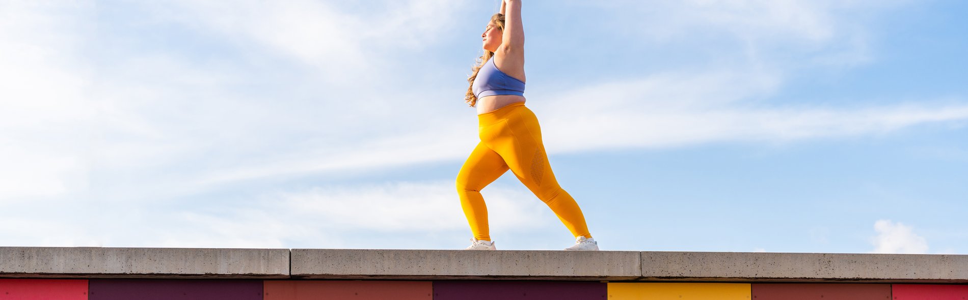 Frau in gelber Leggings und blauem Sporttop macht Yoga auf einer Mauer vor blauem Himmel