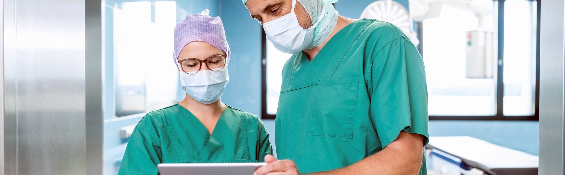 Zwei Ärzte stehen vor dem Operationssaal, schauen auf ein Tablet und besprechen die OP