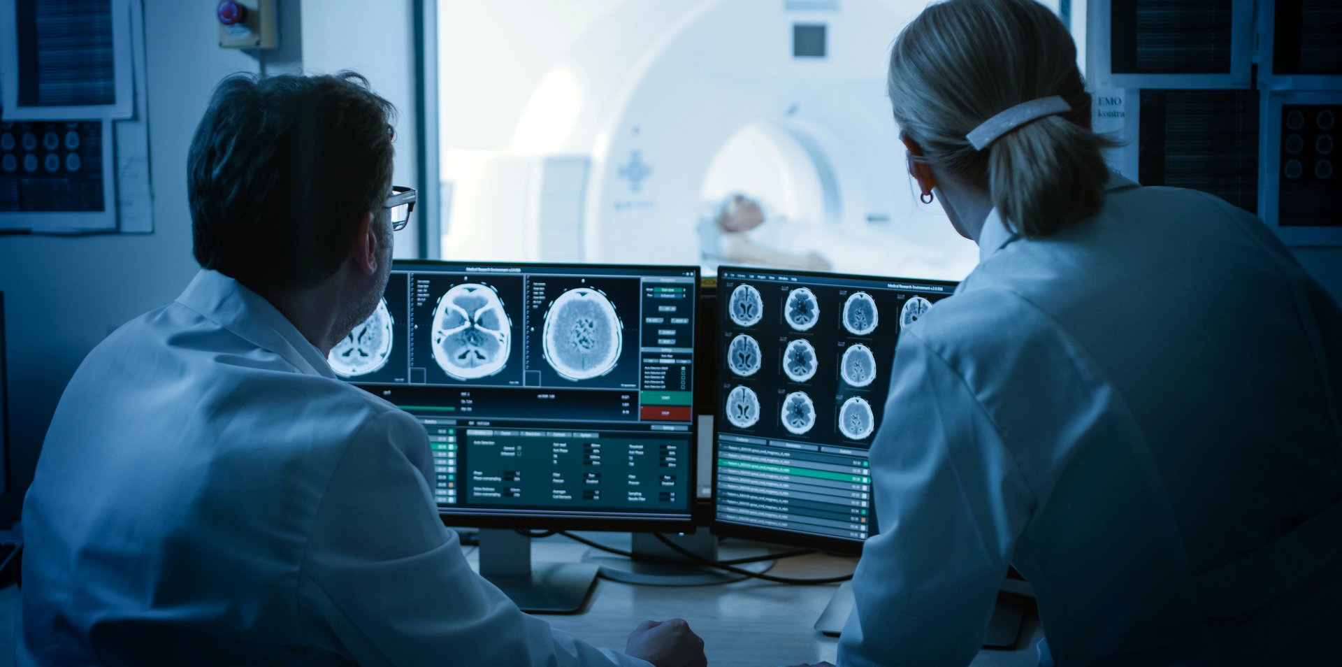 Zwei Ärzte der Neurologie untersuchen Gehirnscans auf dem Bildschirm. Im Hintergrund ist ein Patient im MRT zu sehen.