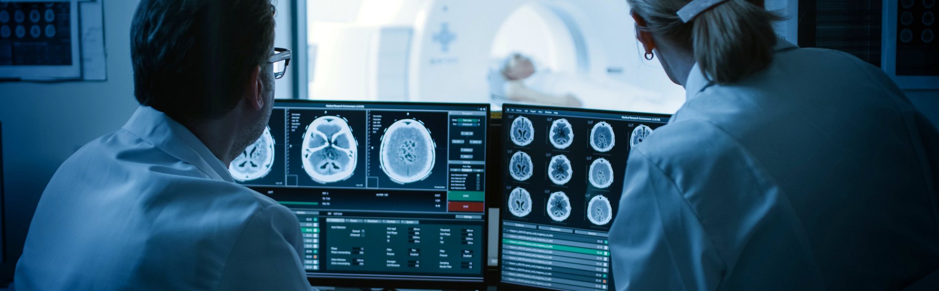 Два врача-невролога изучают снимки мозга на экране. На заднем плане виден пациент на МРТ.